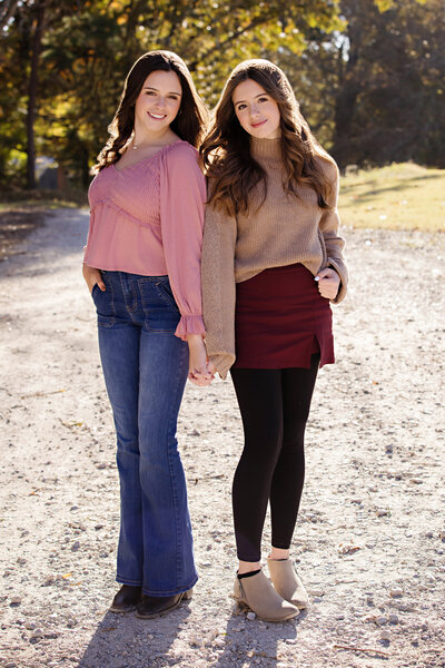 Two sisters standing back to back on gravel roadway framed by colorful fall foliage at Anne Springs Close Greenway Charlotte NC