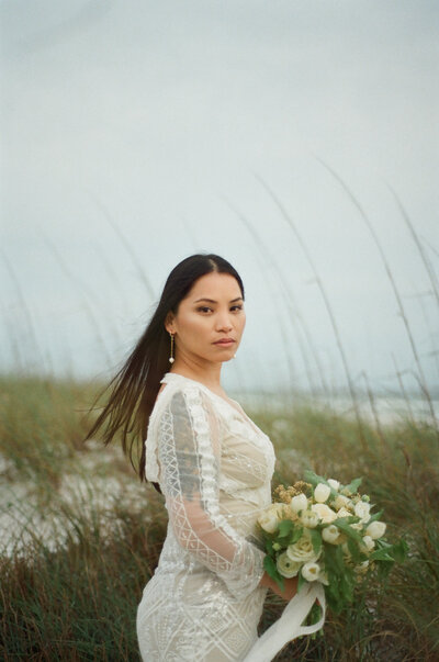 brunette bride poses on beach with white & green bridal bouquet and hair blowing.  Portrait taken by Brittney Stanley of Be Seen Photos - panama city fl photographer