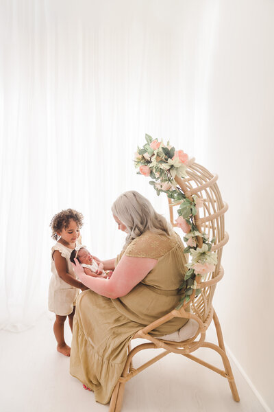 A tender moment as a mother holding a newborn interacts with her curious toddler in a softly lit room, evoking a sense of warmth, family, and love. Taken by Twin Cities Newborn Photographer, Fig and Olive Photography