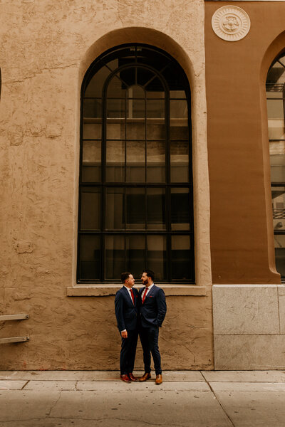 two grooms standing together in front of a large window in downtown Albuquerque