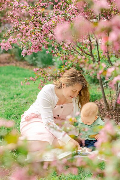 Mother wearing white cardigan and light pink dress playing with her baby son under pink flowering trees. Photos by Chicago Family Photographer Kristen Hazelton