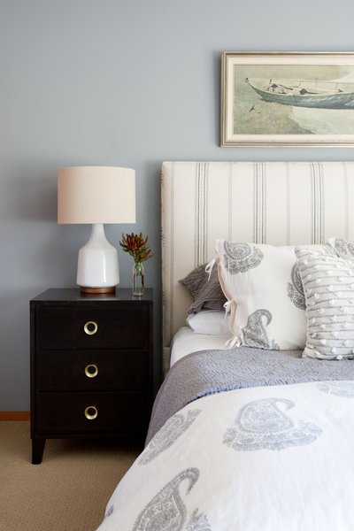 Striped upholstered bed with cream and gray paisley bedding, black dresser with brass hardware, and vintage oil painting of boat on a gray wall