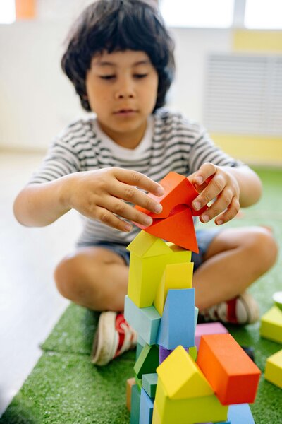 A child in a striped shirt is carefully stacking colorful foam blocks into a tall tower while sitting on a green carpet.