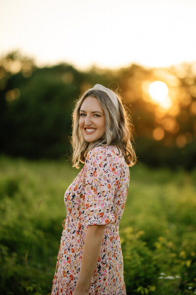 gatlinburg photographer headshot with woman standing in a spring field in the Smoky Mountains with the sun shining through the trees in the distance at sunset 