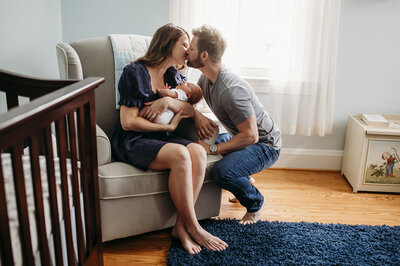 Woman in blue dress sitting in nursery rocking chair holding newborn kissing man in grey shirt and blue jeans near Baltimore Maryland