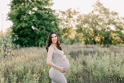expecting mom wearing a beige dress holds belly while smiling in a green field.