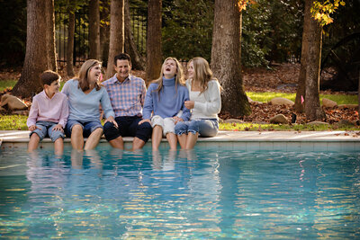 A family of five with parents, two teenage girl and tween son sitting on the edge of the pool dipping their feet and everyone is laughing hard.