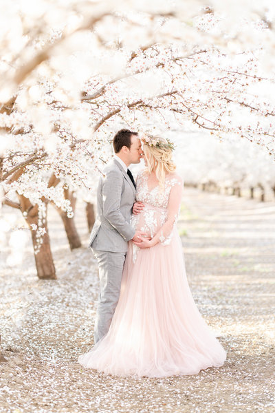 A maternity photography session taken by Bay area photographer shows an expecting mother caressing her baby bump in a field of almond blossoms with her partner.