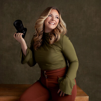 The owner of Kara Reese Photography, Kara Krause, is pictured with her camera. She is wearing a white shirt and has a brown hat.