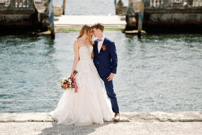 Bride and groom stand  in front of turquoise waters with their foreheads touching.