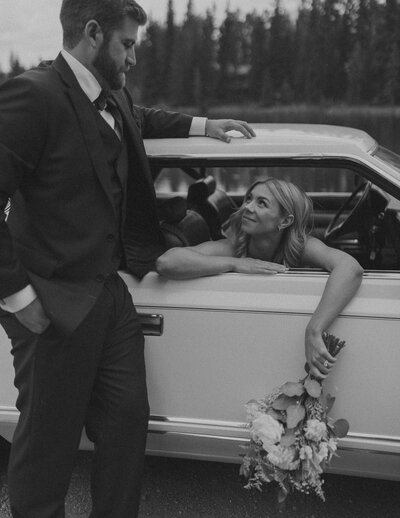 groom leaning against white vintage car while bride leans out the window