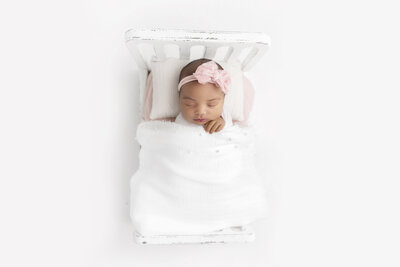 A newborn baby girl sleeps in a pink bow in a tiny wooden bed