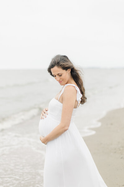 Pregnant woman in flowing white dress embraces her belly during maternity portrait session on Sherwood Island beach