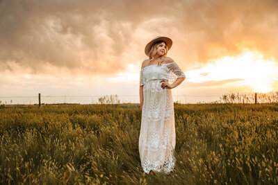 woman in white dress with flat brim hat in field looking off into the sunset
