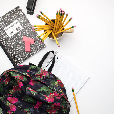 black and floral backpack surrounded by a black composition notebook, two pink erasers, one black stapler, and a white mug filled with yellow number 2 pencils