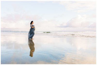pregnant woman standing on beach with reflection showing on the sand below her