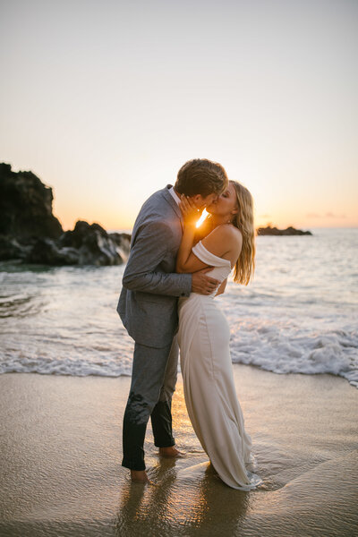 Bride and groom share a kiss as they elope in Waimea Valley. They are standing barefoot on the beach as the waters coming in.