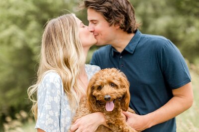 Woman and fiancee kiss while holding their dog during Minneapolis engagement photography session.