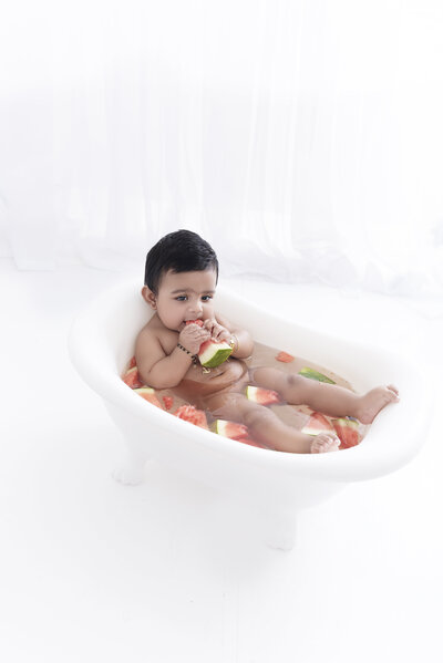 A young toddler baby lays in a tiny bathtub filled with watermelon slices while eating one