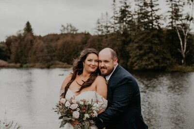 Bride and groom stand in front of a pond at Luso Valley Estate Farm in Orangeville, ON for a late-fall wedding. The bride's back is against her groom's chest, their cheeks are touching and both smiling at the camera. Captured by London, ON-based top wedding photographer Ashlee Ellison.