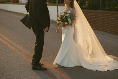Man in tuxedo and women in a wedding dress and long vail holding bouquet of flowers staring at each other with soft smiles in the middle of the street