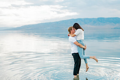 Man is standing in a lake with mountains in the distance, bum lifting up a woman and almost kissing her