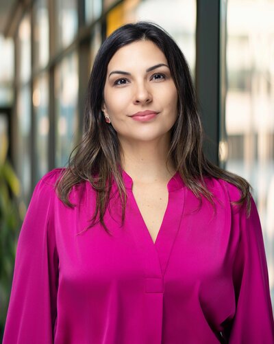 A person stands on stone steps in front of a large stone building, holding a bright red coat. They are wearing a white dress and red shoes. Trees are visible in the background.