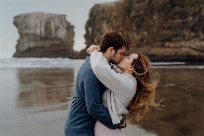 A couple wearing a blue shirt and a pink skirt standing in the water during an engagement photoshoot in Muriwai, Auckland, photographed by waikato photographer haley adele photography