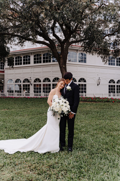 Bride smiles and rests her head on grooms shoulder as they pose in front of beautiful venue. Bride is holding a large bouquet of flowers. Photo taken by Orlando Wedding Photographer Four Loves