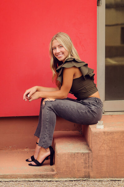 Raegan is sitting on a step in a green top with a ruffled sleeve in front of a red building.