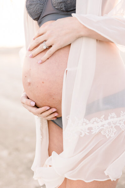 Expecting mother's bare belly with her hands gently holding baby bump, white lace robe blowing in the wind, taken by sacramento family photographer Kelsey Krall