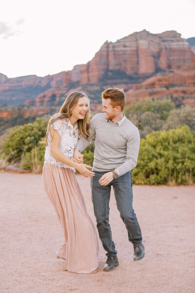 Couple playing tag in Sedona az wearing a white lace shirt and pink skirt and a gray sweater over a button down and jeans