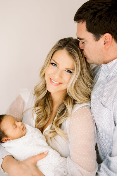 Mom holding newborn baby boy in white blanket smiles at camera while dad kisses her head