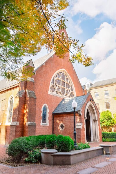 A classic red-brick church with a large circular stained glass window sits under a partly cloudy sky. Greenery surrounds the building, and autumn leaves appear at the top of the image, adding a touch of color.