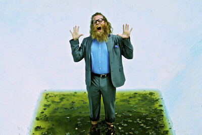 Musician portrait Ben Caplan standing in puddle in drained pool holding up his arms and screaming