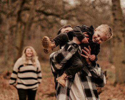 laughing and smiling father chucking his son up in the air as the mom watches them in the distance at a family photoshoot in the west midlands in a woodland location