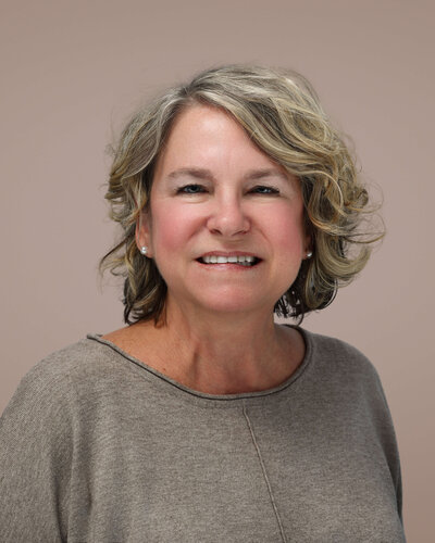 A person stands on stone steps in front of a large stone building, holding a bright red coat. They are wearing a white dress and red shoes. Trees are visible in the background.