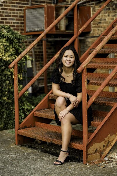 A senior girl in a black dress leans sits on a red metal staircase during her senior photo session