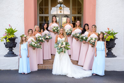 Bride with her bridesmaids in blush pink dresses and flowers pose on the stairs at Ravisloe Country Club.