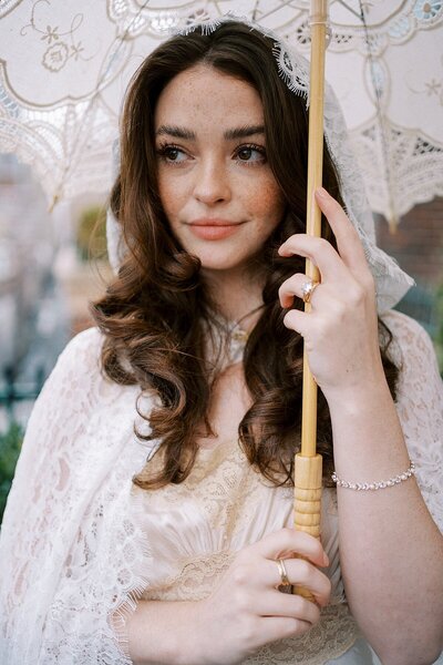 Bride standing in rain with umbrella in a vintage wedding gown with white lace umbrella