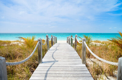 A wooden boardwalk with rope railings leads through grassy dunes to a tranquil turquoise sea under a blue sky with scattered clouds. Palm fronds are visible on either side of the path.