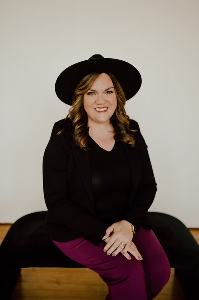 Woman in a black hat and blazer, smiling while seated on a black bench, with her hands on her crossed knee, perhaps contemplating stress-free wedding plans.