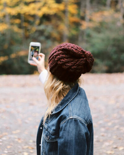 Woman in a blue jacket and red beanie who has her arm outstretched, recording a Tiktok video