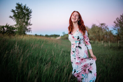 Caucasian redhead wearing long flowing dress in tall grass at sunset and laughing