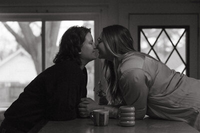 An engaged couple sits across one another in their kitchen, leaning in for a suspenseful kiss
