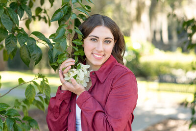 High school senior girl holding whit flowers hanging off of a tree smiling at the camera.