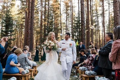 A blond bride wearing a mermaid cream color gown laughs and smiles at her groom wearing his Coast Guard white uniform at paper airplanes are tossed down the aisle for their intimate wedding at Five Pine Lodge in Sisters, Oregon. | Erica Swantek Photography