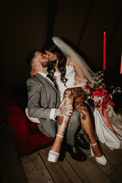 A flash photo of a tattooed bride and groom kiss on the sofa at their wedding day. 