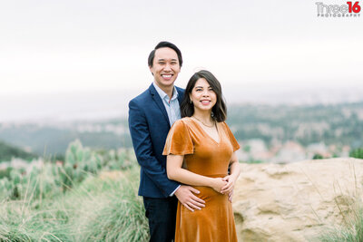 Groom to be holds his fiance from behind as they pose for engagement photos atop a hill in Coastal Vista View Park in Newport Beach
