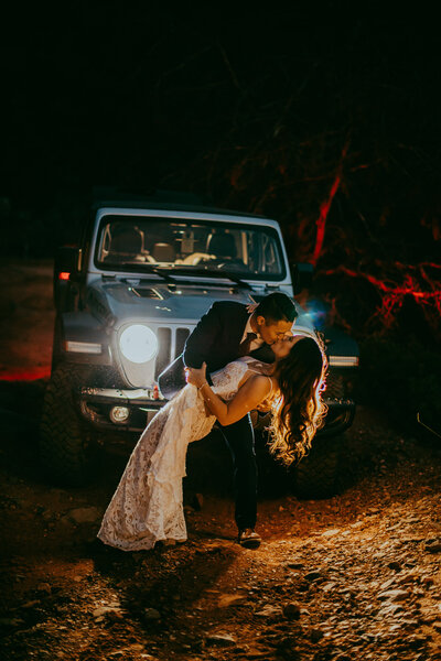 man and woman kiss in front of jeep headlights
